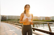 © maxbelchenko - An athletic woman wearing wireless headphones runs across a bridge in the sun's rays. A young woman in athletic clothes enjoying the cityscape on a sunny day. Concepts of running, sports, and health.