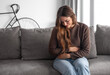 © luciano - Young and beautiful sick woman sits at home clutching her aching stomach, suffering from abdominal discomfort caused by indigestion, gastritis, menstrual cramps, or digestive problems, cramps