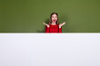 © deagreez - Young woman in red shirt stands behind a white panel with a green background smiling wide and raising hands in a joyful promotional lifestyle fashion moment