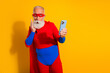 © deagreez - Elderly bearded man in a red and blue superhero costume takes a selfie with a blue phone against a bright yellow background at a playful event