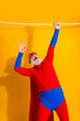 © deagreez - Energetic senior superhero performer hangs from a rope in bold red and blue with cape against a bright yellow background for festive party and advertising imagery