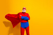 © deagreez - Senior bearded man in red superman costume with blue cape stands confident against a bright yellow background for bold entertainment imagery