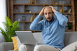 © Liubomir - Young man holding his head in despair, feeling stressed and shocked after seeing bad news on the laptop screen, experiencing frustration and worry while sitting on the sofa