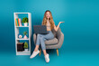 © deagreez - Young chic woman with laptop seated in a cozy chair beside a white bookshelf in a bright blue studio