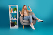 © deagreez - Young stylish woman relaxing on a modern chair with shelf decor in a blue studio
