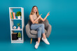 © deagreez - Young woman sits in grey armchair beside a white shelf against a blue background and smiles while pointing to the side