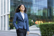 © Liubomir - Confident young businesswoman smiling, walking outdoors with hand in pocket, against a backdrop of modern glass office buildings, representing professional success and urban career