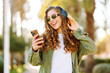 © maxbelchenko - Portrait of young woman wearing wireless headphones listening to music while walking in the park. Happy woman in sunglasses holds smartphone and enjoys sunny day. Concept of walk, music and outdoor.