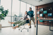 © deagreez - Businesswoman enjoys a playful moment as a colleague pushes her chair in a modern busy office