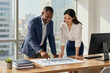 © f_bossa - Two professionals in business attire collaborate over a printed bar chart on a wooden desk in a bright office with large windows. They point at data while smiling, with city buildings visible outside.