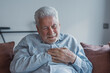 © Daniel - A senior man sits on a sofa at home holding his chest in pain, showing symptoms of a heart attack while experiencing distress and urgent health concerns in a domestic setting
