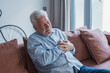 © Daniel - A retired man sits alone on a sofa pressing his chest with a pained expression, reflecting a possible heart attack and the frightening reality of sudden health problems