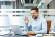 © Liubomir - Smiling man engaging in a virtual meeting, communicating remotely from an office setting, actively using his laptop for online business networking and distance learning