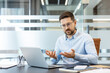 © Liubomir - Young businessman in a light blue shirt and glasses gesturing with his hands while participating in an online video conference, or remote meeting using a laptop in a modern office