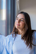 © Daniel - Standing near a window, a young woman gazes at the winter scenery as soft sunlight reflects on her happy face, leaving copy space for text in a serene and bright home interior