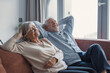 © Daniel - A grey-haired husband and wife sit side by side on a sofa with hands behind their heads, daydreaming together and embracing relaxation and emotional balance at home