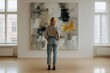 © VICHIZH - Woman with curly hair wearing striped shirt and jeans standing in bright gallery room looking at large abstract painting on white wall between two windows.