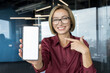 © Liubomir - Happy young businesswoman showing a smartphone with an empty white screen for mock up while presenting an application or platform, in a modern corporate office environment