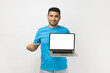 © khosrork - Portrait of pleased positive unshaven man wearing blue T- shirt standing standing holding laptop, pointing at empty display of notebook. Indoor studio shot isolated on gray background.