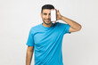 © khosrork - Smiling happy attractive unshaven man wearing blue T- shirt standing covering his eye with smart phone with blank white screen, advertisement area. Indoor studio shot isolated on gray background.