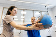 © wedmoments.stock - Physiotherapist assisting male patient with rehabilitation exercises using a weighted medicine ball in a bright, modern therapy room with exercise equipment visible