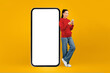 © Prostock-studio - A young woman stands next to a large blank phone display. She is looking at her smartphone and smiling. The background is bright yellow, creating a lively atmosphere.