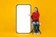 © Prostock-studio - A woman in a red hoodie sits on a chair and points towards a blank screen. The background is yellow, and she looks happy while promoting something. The scene has a playful and inviting feel.