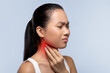 © Prostock-studio - A woman stands in an indoor space, holding her neck with a pained expression. She appears to be experiencing discomfort or pain in her throat area, possibly due to an illness.