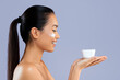 © Prostock-studio - A woman is smiling while applying skincare cream to her face. She holds a small bowl in one hand. The setting has a plain background and good lighting. This shows a moment of self-care.
