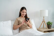 © Kawee - Asian young woman chatting on mobile phone while sitting on bed at home.