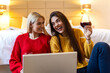 © Graphicroyalty - Happy women drinking red wine and using laptop in bedroom. Friendship, technology and leisure concept. Two female friends laughing while watching content on a computer screen at home or in hotel.