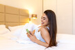 © Graphicroyalty - Cheerful woman using smartphone while lying in bed. Social media and communication concept. Beautiful brunette female smiling at mobile phone screen while relaxing under white duvet in bedroom.
