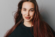 © SHOTPRIME STUDIO - Young woman with long hair and natural makeup smiling at camera on gray background. Female portrait with casual black top and confident expression for beauty and lifestyle.