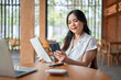 © bongkarn - Pretty asian woman holding book looking at phone aside laptop while sitting at wooden table in cafe.