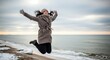 © Erisna Yolanda - A woman in a long coat jumps on a snowy beach with her arms outstretched towards a cloudy sky and ocean
