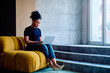 © we.bond.creations - Concentrating businesswoman using a laptop while sitting on a yellow designer sofa in a modern office space, remote work concept
