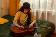 © _KUBE_ - Caucasian female student practicing guitar indoors while sitting on bed. Home music education scene. Artistic development and self improvement. Wide shot