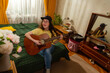 © _KUBE_ - Happy young woman plays guitar in a home setting developing musical skills and creativity surrounded by plants and calm domestic atmosphere.