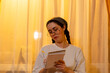 © _KUBE_ - Attractive young woman wearing eyeglasses keeping a journal at home during golden hour. Calm evening routine and mindfulness practice. Concept of mental health, and productivity in modern lifestyle.