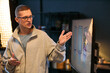 © DragonImages - Caucasian young adult man gesturing while explaining information on large monitor during presentation, standing in modern office environment with focused expression