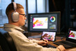 © DragonImages - Caucasian young adult man wearing headphones using digital pen tablet while editing photo on computer monitor and tablet screen in modern workspace, focusing on creative work