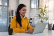 © MINAE - Asian woman smiling and typing on a laptop computer while sitting at a bright desk, representing remote work, productivity, and digital communication in a professional environment