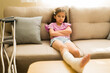 © AntonioDiaz - Preteen girl sitting on a couch with a broken leg in a cast, looking unhappy and frustrated with her injury