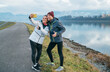 © Soloviova Liudmyla - Happy young active couple in athletic jackets and beanies taking a selfie with a smartphone and giving a thumbs up on a paved riverside path during a long run day training session on an overcast day