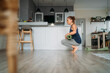 © Soloviova Liudmyla - Healthy young woman crouching on a wooden floor while mindfully rolling up her green yoga mat after completing a morning wellness routine in a bright and spacious modern home kitchen and living area