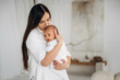 © standret - Mother holding newborn baby in white clothing, softly gazing at child in a bright, minimalistic indoor setting with neutral tones and soft textures
