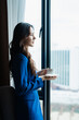 © Tj - Young woman in a blue suit smiling, holding a white cup and saucer, and looking out a large window at a blurred cityscape, contemplating the urban environment