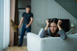 © Tj - Upset woman feeling stressed and frustrated, lying on a sofa while a man is seen in the background holding a broom, indicating a challenging relationship or conflict at home