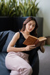 © Tj - Woman sitting comfortably on a sofa, engrossed in reading a book, finding a moment of peace and relaxation in her lifestyle, surrounded by lush indoor greenery in a quiet setting