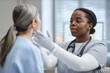 © Seventyfour - Black young adult woman doctor examining face of Caucasian middle aged woman patient in medical office, wearing gloves and stethoscope, focusing on patient during checkup